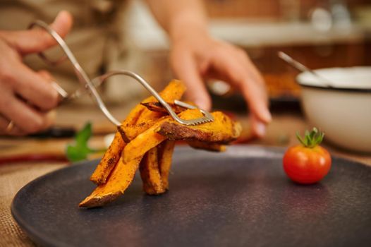 Close-up of the hand of female chef serving a dish, putting roasted organic wedges of sweet potato on a ceramic plate with one ripe cherry tomato. Restaurant menu. Delicious vegan meal. Healthy eating