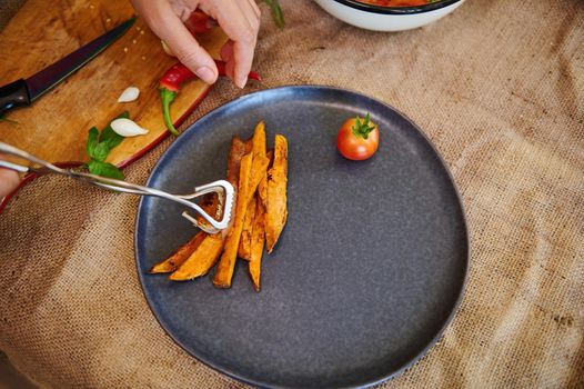 View from above of a chef's hand serving plate, putting sweet potato fries on a dark ceramic plate. Copy advertising space. Web banner. Menu for restaurants. Cooking master class. Healthy vegan food