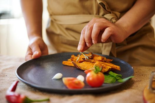 Close-up of serving dish: a chef puts a rosemary leaf on the top of roasted wedges of organic batata, slicing cherry tomato on a wooden chopping board and ornate the sweet potato.