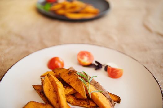 Close-up of a plate with slices of fried organic sweet potatoes - batata drizzled with olive oil and rosemary in a table with linen tablecloth. Healthy vegetarian food. Vegan's Day. Restaurant menu