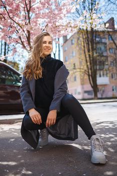 A young slender female model with long wavy hair and, dressed in a gray coat, sneakers, sits on the street. Spring blossom trees woman girl laugh and rejoice