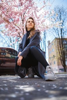 A young slender female model with long wavy hair and, dressed in a gray coat, sneakers, sits on the street. Spring blossom trees woman girl laugh and rejoice