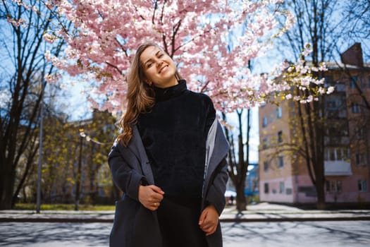 A young slender female model with long wavy hair and, dressed in a gray coat, sneakers, stands on the street near a flowering shrub with beautiful pink flowers in the background and poses.
