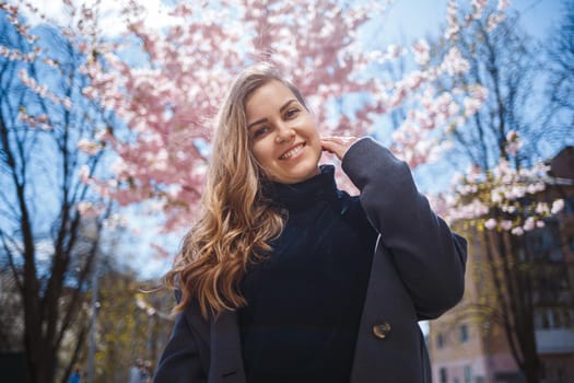A young slender female model with long wavy hair and, dressed in a gray coat, sneakers, stands on the street near a flowering shrub with beautiful pink flowers in the background and poses.