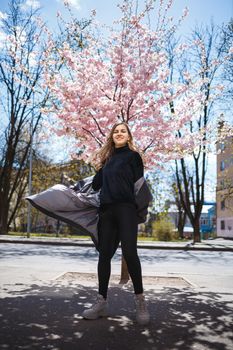 Young slender female model with long wavy hair and, dressed in a gray coat, sneakers, spinning on the street. Spring blossom trees woman girl laughs and runs to rejoice