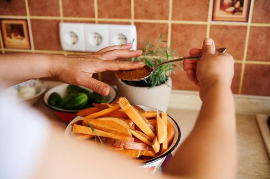 Close-up. Selective focus on hands of housewife holding spoon with culinary spices and sprinkling them into bowl with sweet potato wedges while seasoning dishes with culinary herbs