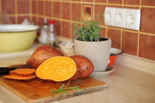 Selective focus on whole and sliced tuber of sweet potato, batata, a kitchen knife on a wooden cutting board against cultivated rosemary in ceramic pot for meal seasoning on a beige kitchen countertop