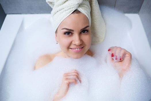 Girl woman with a white towel on her head lies in the bathroom in a white bath. Foam for around. Relax after a hard day. Spa relaxing treatment
