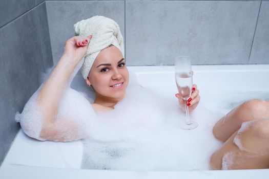 Girl woman with a white towel on her head lies with a glass of champagne in a white bathtub. In her hands a lot of pee soap. Full whirlpool foam. Relax after a hard day. Spa relaxing procedure