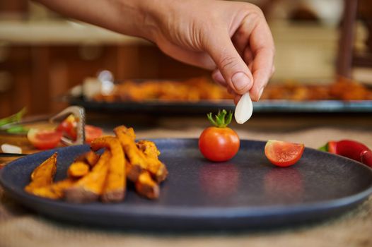 Roasted sweet potatoes. Baked dish with organic batata with cherry tomatoes and rosemary herbs. Banner. menu, Recipe. Close-up chef's hand putting a garlic clove on a plate with fried sweet potatoes
