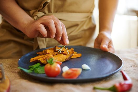 Close-up of the hands of a housewife or chef decorating dishes of fried sweet potato with cherry tomatoes and herbs, preparing delicious healthy vegan meal for lunch. Selective focus