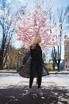 Young slender female model with long wavy hair and, dressed in a gray coat, sneakers, spinning on the street. Spring blossom trees woman girl laughs and runs to rejoice