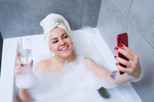 A beautiful woman with a towel on her head lies with a glass of champagne in a white bath and takes a selfie on the phone. Relax after a hard day. Relaxing spa treatment