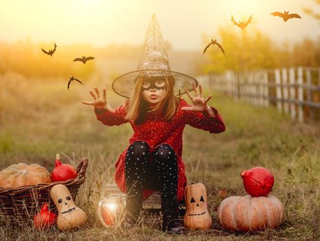 Little girl in halloween costume sitting next to pumpkins while frightening at camera on autumn nature
