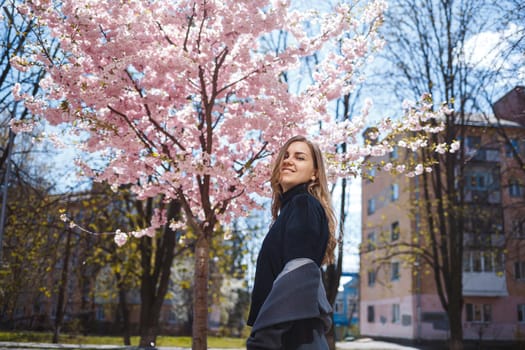 A young slender female model with long wavy hair and, dressed in a gray coat, sneakers, stands on the street near a flowering shrub with beautiful pink flowers in the background and poses.