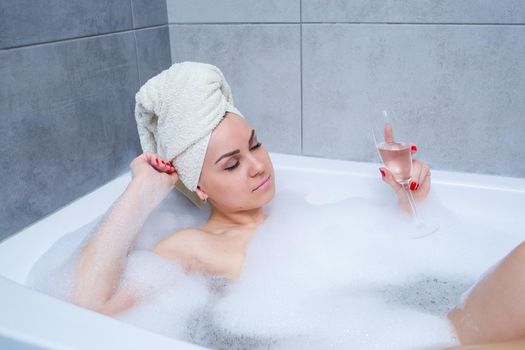 Girl woman with a white towel on her head lies with a glass of champagne in a white bathtub. In her hands a lot of pee soap. Full whirlpool foam. Relax after a hard day. Spa relaxing procedure