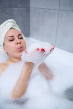 Girl woman with a white towel on her head lies in the bathroom in a white bath. In the hands of a lot of muddy soap. Full whirlpool foam. Relax after a hard day. Spa relaxing treatment