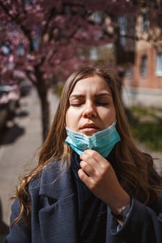 Girl, young woman in a protective sterile medical mask on her face, looking at the camera outdoors, in the spring garden. Air pollution, virus, pandemic coronavirus concept.