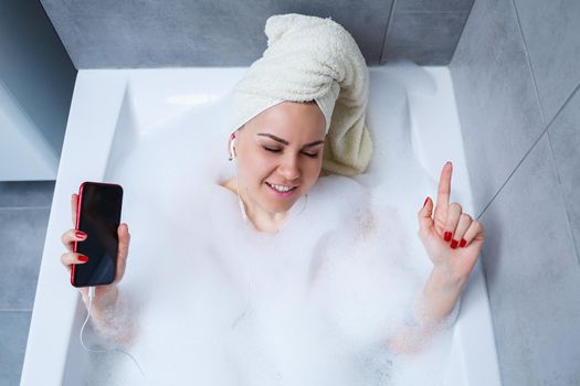Beautiful young woman listening to music and singing while taking a bath at home. Relax after a hard day. Spa relaxing procedure.