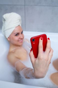 Girl woman with a towel on her head lies with a glass of champagne in a white bathtub and takes a selfie on the phone. In her hands a lot of drunk soap. Relax after a hard day. Spa relaxing procedure