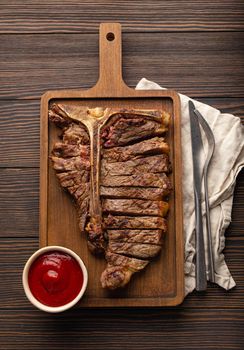Sliced grilled medium rare meat beef steak T-bone barbecue with red tomato sauce served on wooden cutting board and textile napkin from above with fork and knife