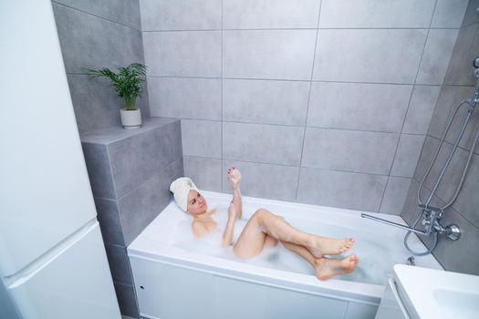 Girl woman with a white towel on her head lies in the bathroom in a white bath. In the hands of a lot of muddy soap. Full whirlpool foam. Relax after a hard day. Spa relaxing treatment