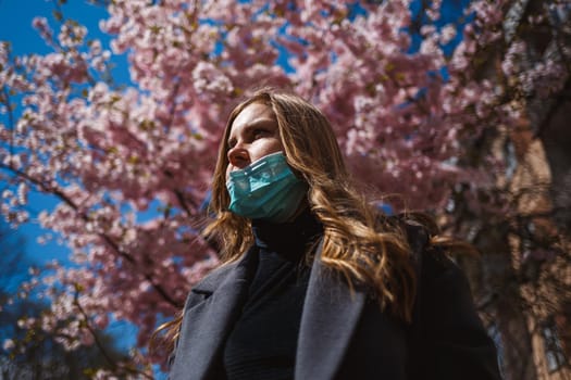 Girl, young woman in a protective sterile medical mask on her face, looking at the camera outdoors, in the spring garden. Air pollution, virus, pandemic coronavirus concept.