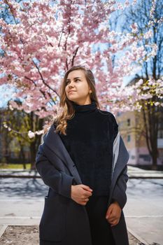 A young slender female model with long wavy hair and, dressed in a gray coat, sneakers, stands on the street near a flowering shrub with beautiful pink flowers in the background and poses.