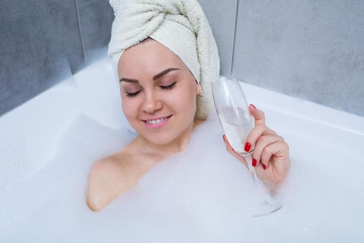 Girl woman with a white towel on her head lies with a glass of champagne in a white bathtub. In her hands a lot of pee soap. Full whirlpool foam. Relax after a hard day. Spa relaxing procedure