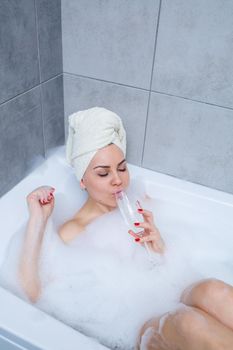 Girl woman with a white towel on her head lies with a glass of champagne in a white bathtub. In her hands a lot of pee soap. Full whirlpool foam. Relax after a hard day. Spa relaxing procedure