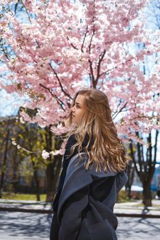 Young slender female model with long wavy hair and, dressed in a gray coat, sneakers, spinning on the street. Spring blossom trees woman girl laughs and runs to rejoice