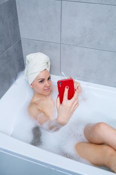 Girl woman with a towel on her head lies with a glass of champagne in a white bathtub and takes a selfie on the phone. In her hands a lot of drunk soap. Relax after a hard day. Spa relaxing procedure