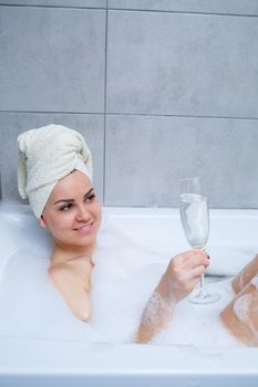 Girl woman with a white towel on her head lies with a glass of champagne in a white bathtub. In her hands a lot of pee soap. Full whirlpool foam. Relax after a hard day. Spa relaxing procedure