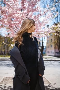 Young slender female model with long wavy hair and, dressed in a gray coat, sneakers, spinning on the street. Spring blossom trees woman girl laughs and runs to rejoice
