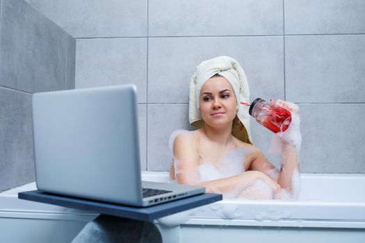 A young woman with a towel on her head drinks juice watching a movie on a laptop while sitting in a bath in a beauty salon. Relax in the bathroom without linen. Body care and relaxation concept.