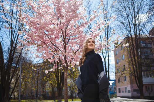 A young slender female model with long wavy hair and, dressed in a gray coat, sneakers, stands on the street near a flowering shrub with beautiful pink flowers in the background and poses.