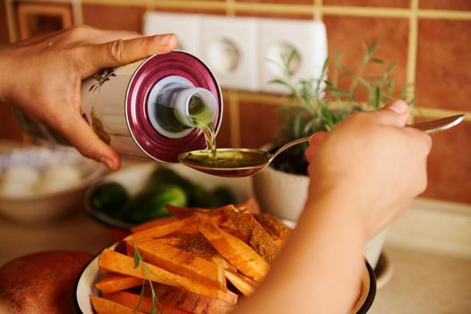 Selective focus. Close-up of chef's hands pouring organic olive oil on a spoon, and sprinkling it over organic sweet potato slices while preparing lunch in the home kitchen