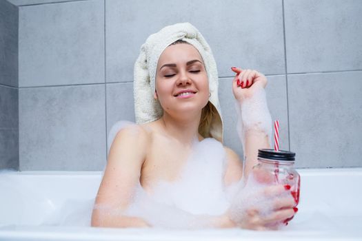 Beautiful young woman with a towel on her head drinking a cocktail takes a bath at home. Relax after a hard day. The spa is a relaxing procedure.