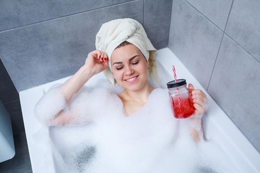 Beautiful young woman with a towel on her head drinking a cocktail takes a bath at home. Relax after a hard day. The spa is a relaxing procedure.