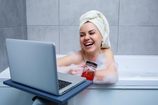 A young woman with a towel on her head drinks juice watching a movie on a laptop while sitting in a bath in a beauty salon. Relax in the bathroom without linen. Body care and relaxation concept.