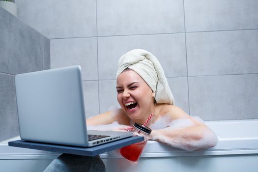 A young woman with a towel on her head drinks juice watching a movie on a laptop while sitting in a bath in a beauty salon. Relax in the bathroom without linen. Body care and relaxation concept.