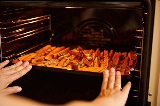 Close-up of chef's hands, putting a baking sheet with raw organic sweet potatoes wedges, seasoned with culinary herbs and spices and sprinkled with olive oil, into an oven while cooking vegan dinner