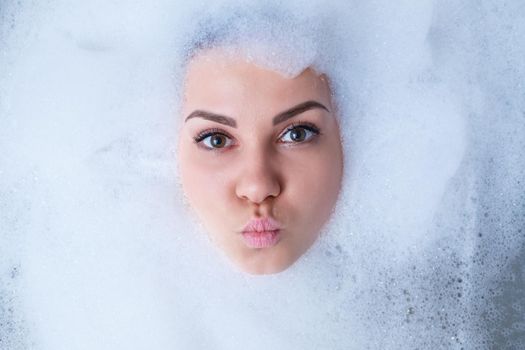 Closeup portrait of a girl in a bathtub and white foam around her face. Different emotions, grimaces, woman smile