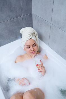 Girl woman with a white towel on her head lies with a glass of champagne in a white bathtub. In her hands a lot of pee soap. Full whirlpool foam. Relax after a hard day. Spa relaxing procedure