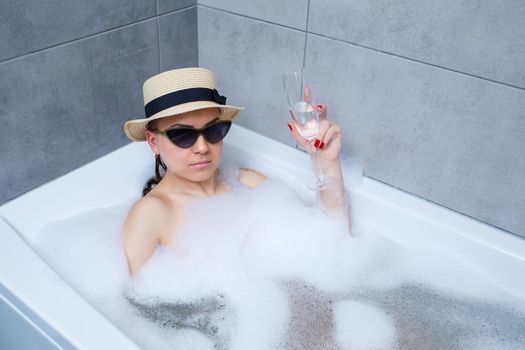 Beautiful young woman in a hat on her head and sunglasses drinking a cocktail takes a bath at home. Relax after a hard day. The spa is a relaxing procedure.