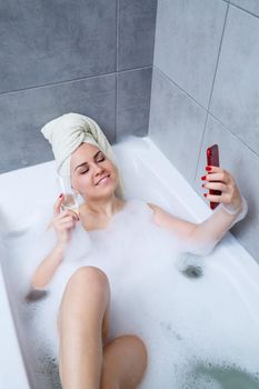 A beautiful woman with a towel on her head lies with a glass of champagne in a white bath and takes a selfie on the phone. Relax after a hard day. Relaxing spa treatment