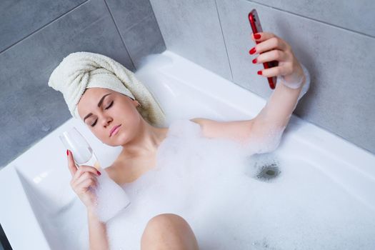 A beautiful woman with a towel on her head lies with a glass of champagne in a white bath and takes a selfie on the phone. Relax after a hard day. Relaxing spa treatment