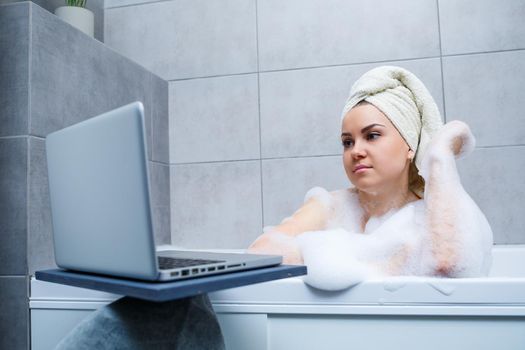 A young woman with a towel on her head is watching a movie on a laptop while sitting in a bath in a beauty salon. Relax in the bathroom without linen. Body care and relaxation concept.