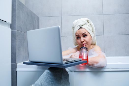 A young woman with a towel on her head drinks juice watching a movie on a laptop while sitting in a bath in a beauty salon. Relax in the bathroom without linen. Body care and relaxation concept.