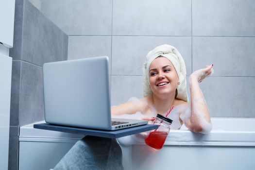 A young woman with a towel on her head drinks juice watching a movie on a laptop while sitting in a bath in a beauty salon. Relax in the bathroom without linen. Body care and relaxation concept.